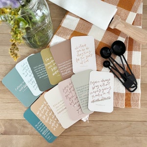May include: A collection of inspirational quote cards in various pastel colors, featuring handwritten-style text. The cards are arranged on a checkered orange and white cloth, with black measuring spoons and a glass jar with flowers.
