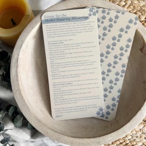 May include: Two prayer cards with text, "Litany for the Mother Enduring Miscarriage," rest in a wooden bowl. One card features the litany, the other has a pattern of small blue flowers. A lit candle is in the background.
