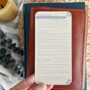 May include: A brown leather wallet with a card that reads "Litany for the Mother Enduring Miscarriage." The card has a floral design and text in a script font. The wallet is on top of a blue book.