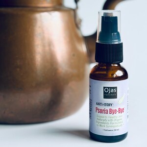 May include: A brown glass bottle with a black pump and a white label. The label reads "Ojas naturals ANTI-ITCHY Psoria Bye-Bye" and "1 fluid ounce (30 ml)". A copper kettle is in the background.