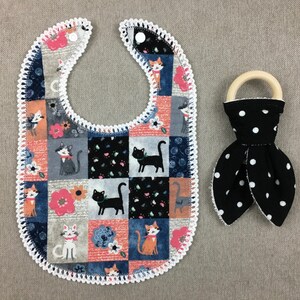 May include: A patchwork baby bib with a crocheted edge and a black and white polka dot teether with a wooden ring. The bib features a cat pattern with orange, blue, gray, and pink colors.