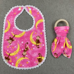 May include: Pink baby bib with a white crocheted edge and a moon and monkey print. A matching pink and white fabric teether with a wooden ring.