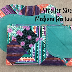 May include: A teal and purple patterned baby blanket with a teal backing. The blanket is folded in half and has a rounded edge. The text "Stroller Size Medium Rectangle" is printed on the blanket.