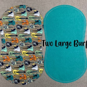 May include: Two large, oval-shaped burp cloths. One is turquoise blue and the other has a pattern of colorful guitars on a beige background. The text "Two Large Burps" is printed on the turquoise cloth.