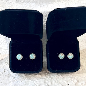 May include: Two pairs of round stud earrings with a light blue and white speckled design, presented in open black velvet jewelry boxes. The earrings are set against a white background.