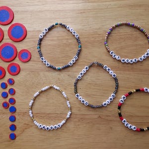 May include: An overhead shot displays several beaded bracelets with white letter beads spelling out phrases like "BOY SHIT", "GET MORE DOWN", and "KEEP IT ROLLIN". Also, there are red and blue circular wooden pieces.