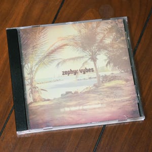 May include: A CD case featuring the album "zephyr vybes." The cover art depicts a tropical beach scene with palm trees, a sandy shore, and the text "zephyr vybes" and "the island of enchantment." The case is clear with a black spine.