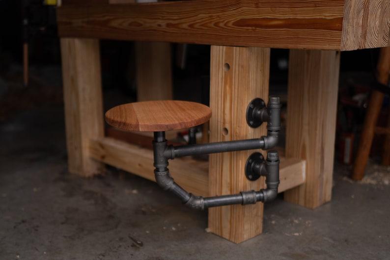 May include: A wooden stool with a black metal frame attached to a wooden workbench. The stool is made of wood and has a round seat.