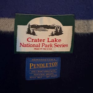 May include: Close-up of a wool blanket with green, blue, and white stripes. The blanket features two rectangular tags. The top tag reads "Crater Lake National Park Series" and "Made in the USA." The bottom tag reads "Pendleton" and "100% Pure Virgin Wool."