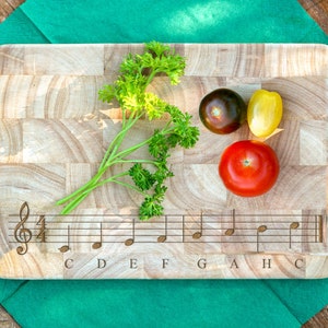 Può includere: Un tagliere in legno con un design a pentagramma musicale e note incise sulla superficie. Il tagliere è di colore marrone chiaro ed è decorato con un rametto di prezzemolo, un pomodoro rosso, un peperone giallo e un pomodoro viola scuro.