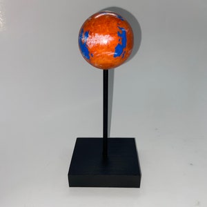 Puede incluir: Un peque&ntilde;o globo terr&aacute;queo naranja y azul sobre un soporte negro. El globo es una representaci&oacute;n de un planeta con nubes blancas y oc&eacute;anos azules.
