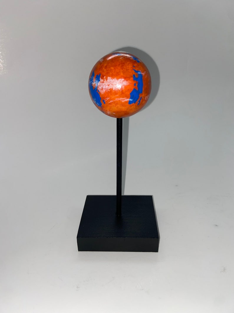 Puede incluir: Un peque&ntilde;o globo terr&aacute;queo naranja y azul sobre un soporte negro. El globo es una representaci&oacute;n de un planeta con nubes blancas y oc&eacute;anos azules.