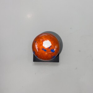 Puede incluir: Una peque&ntilde;a esfera naranja con salpicaduras de pintura blanca y azul. La esfera est&aacute; sentada sobre una base negra.