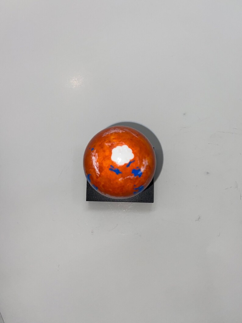 Puede incluir: Una peque&ntilde;a esfera naranja con salpicaduras de pintura blanca y azul. La esfera est&aacute; sentada sobre una base negra.