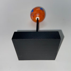 Puede incluir: Un peque&ntilde;o globo terr&aacute;queo naranja con marcas azules se asienta sobre una base cuadrada negra. El globo est&aacute; sobre una fina varilla negra.