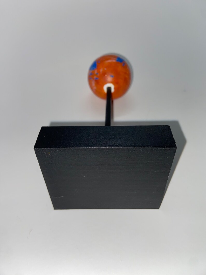 Puede incluir: Un peque&ntilde;o globo terr&aacute;queo naranja con marcas azules se asienta sobre una base cuadrada negra. El globo est&aacute; sobre una fina varilla negra.