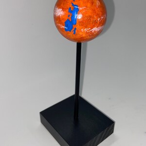 Puede incluir: Una peque&ntilde;a esfera naranja con marcas azules y blancas, que representa a Marte, est&aacute; montada en un soporte negro.