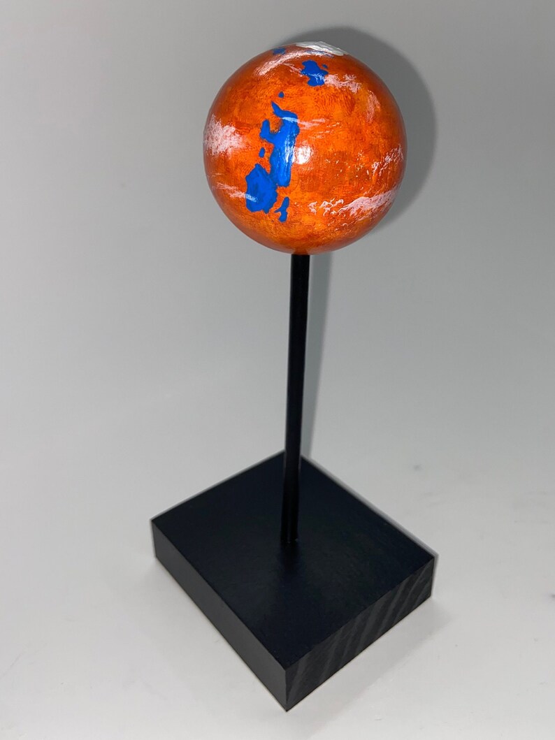 Puede incluir: Una peque&ntilde;a esfera naranja con marcas azules y blancas, que representa a Marte, est&aacute; montada en un soporte negro.