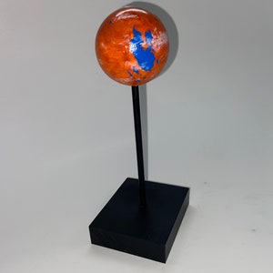 Puede incluir: Un peque&ntilde;o globo terr&aacute;queo naranja y azul sobre un soporte negro. El globo est&aacute; pintado para parecerse a Marte con nubes blancas y agua azul.