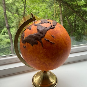 Puede incluir: Un globo decorativo con una cálida combinación de colores naranja y marrón. El globo presenta continentes en un tono marrón más oscuro, montado sobre un soporte de color latón con un meridiano curvo y una base. El fondo muestra una exuberante escena al aire libre verde.