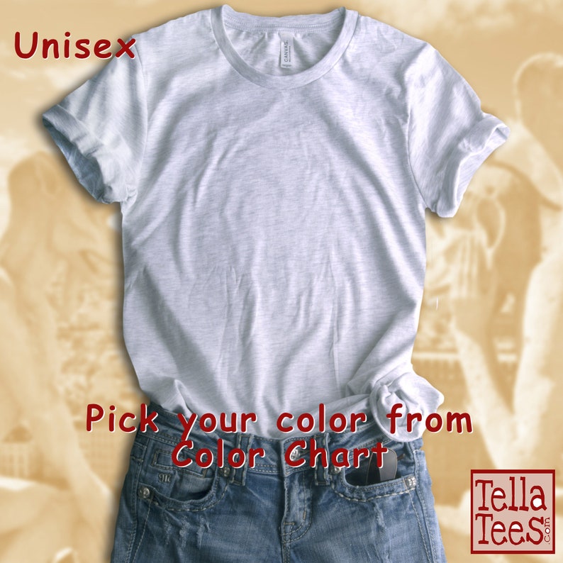 以下が含まれることがあります： ロールアップされた袖のシンプルなライトグレーのユニセックスTシャツ。Tシャツの下部に「Pick your color from Color Chart」というテキストが赤色で印刷されています。Tシャツは青いジーンズの上に置かれています。