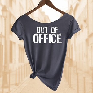 以下が含まれることがあります： 背面に白い文字で「OUT OF OFFICE」とプリントされた濃いグレーのTシャツ。木製のハンガーにかけられています。