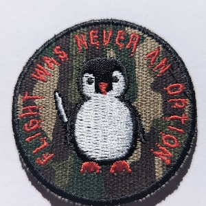 May include: A round, embroidered patch with a camouflage background. A white penguin with a red beak and red feet is holding a knife. The text around the penguin reads "Flight was never an option".
