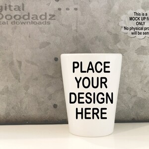Puede incluir: Un vaso de chupito de cerámica blanca con el texto "PLACE YOUR DESIGN HERE" impreso en negro.