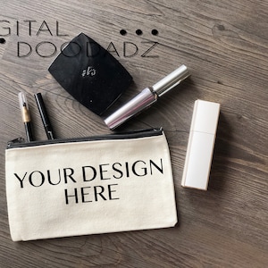 May include: A white canvas zippered pouch with black trim and the text "YOUR DESIGN HERE" printed in black. The pouch is lying on a wood surface with various makeup items around it.