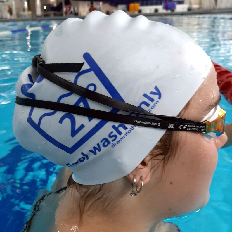 May include: A white swim cap with a blue logo that says "27" and "Speedsocket 2 drawn to wash only.com". The cap is worn by a person in a swimming pool.