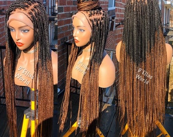 May include: A long, black and brown ombre braided wig with a lace front. The wig is styled with a bun on top. The wig is on a mannequin head and is displayed on a yellow tripod.