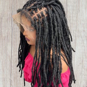 May include: A black dreadlock wig on a mannequin head. The wig features long, individual dreadlocks and a lace front. The mannequin is wearing a pink top. The background is a light brown wooden wall.