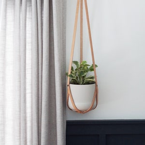 May include: A white ceramic planter with a green and white plant hangs from a light brown leather sling. The sling is attached to a black metal rod. A gray curtain is to the left of the planter.