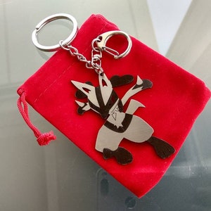 May include: A silver-colored keychain featuring a stylized character design, attached to a chain and keyring. The keychain rests on a small, bright red velvet pouch. The character has black and silver details.
