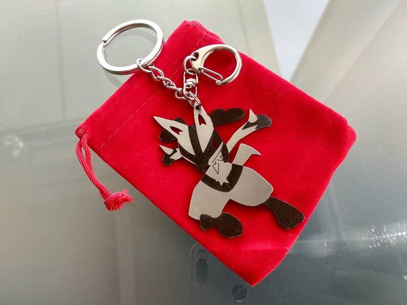 May include: A silver-colored keychain featuring a stylized character design, attached to a chain and keyring. The keychain rests on a small, bright red velvet pouch. The character has black and silver details.
