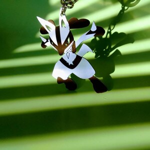 May include: A brown and white Lucario keychain, suspended from a silver chain. The character is in a dynamic pose, with sharp angles and a star-shaped emblem on its chest. The background is a vibrant green with striped shadows.