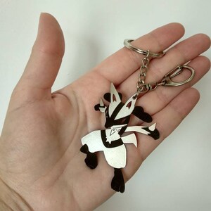 May include: A silver-colored Lucario keychain with black accents. The keychain features a detailed cutout design of the Pok&eacute;mon character, attached to a chain and a keyring. The design is held in a hand.