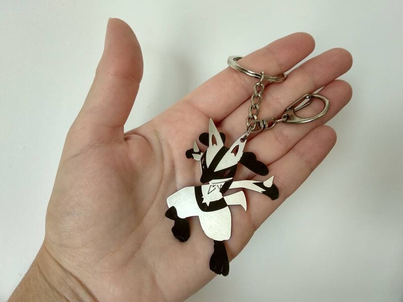 May include: A silver-colored Lucario keychain with black accents. The keychain features a detailed cutout design of the Pok&eacute;mon character, attached to a chain and a keyring. The design is held in a hand.