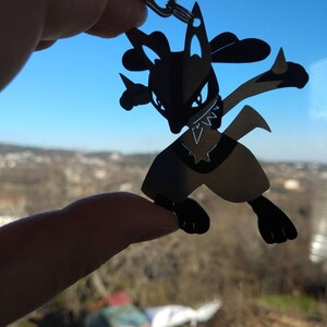May include: Black Lucario keychain. The silhouette of the Pok&eacute;mon character is held up against a bright blue sky and a distant landscape. The keychain is a detailed cutout of the fighting-type Pok&eacute;mon.