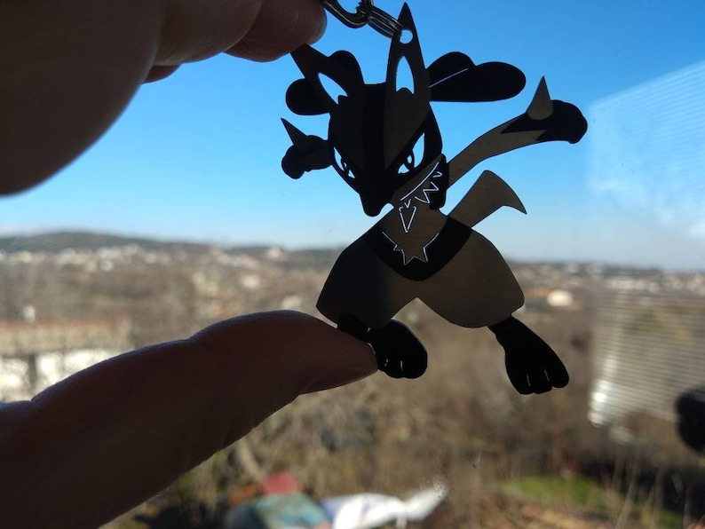 May include: Black Lucario keychain. The silhouette of the Pok&eacute;mon character is held up against a bright blue sky and a distant landscape. The keychain is a detailed cutout of the fighting-type Pok&eacute;mon.