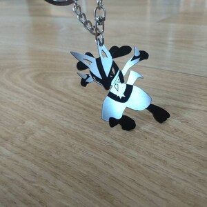 May include: A silver-colored Lucario keychain with black accents, attached to a silver chain. The keychain is in a dynamic pose, with its arms and legs extended. The background is a light brown wooden surface.