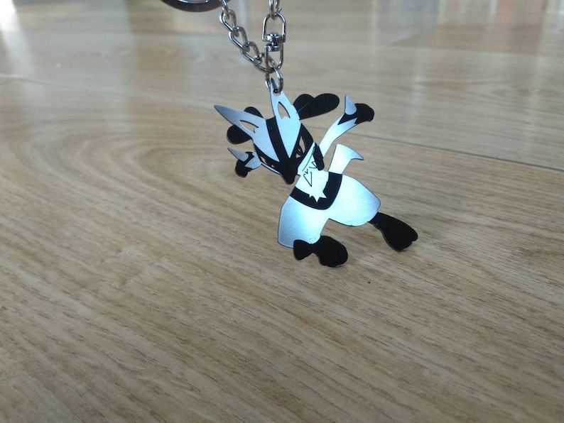 May include: A silver-colored Lucario keychain with black accents, attached to a silver chain. The keychain is in a dynamic pose, with its arms and legs extended. The background is a light brown wooden surface.