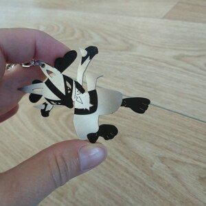 May include: A silver-colored keychain featuring a stylized badger design. The badger has black and white markings and is depicted in a playful pose. The keychain is made of a lightweight material and includes a silver chain and ring for attachment.