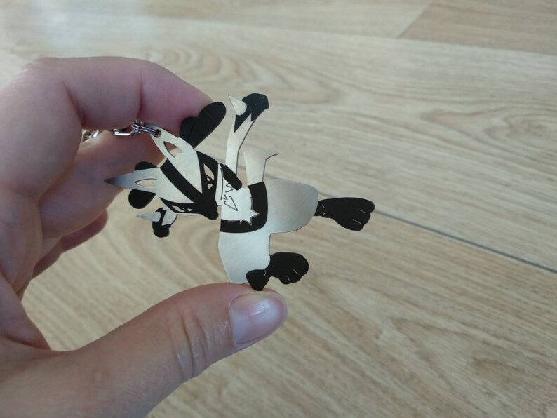 May include: A silver-colored keychain featuring a stylized badger design. The badger has black and white markings and is depicted in a playful pose. The keychain is made of a lightweight material and includes a silver chain and ring for attachment.