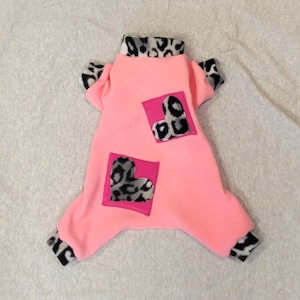 May include: Pink fleece dog pajamas with leopard print hearts. The pajamas have a turtleneck and cuffs.