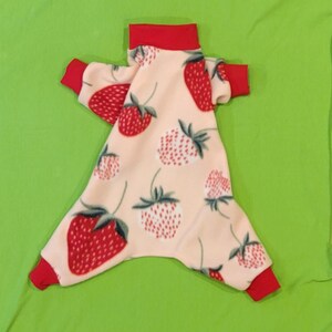 May include: A pink fleece dog jumpsuit with a red collar and a strawberry print.