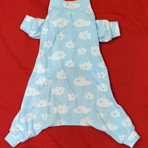 May include: A light blue long-sleeve onesie featuring a repeating pattern of white clouds with smiling faces. The garment has a high neckline and ribbed cuffs at the wrists and ankles, providing a consistent look across the entire piece of clothing.
