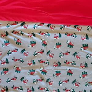 May include: A blue fabric with a repeating pattern of red toy trucks carrying green Christmas trees. The fabric is folded over a bright red fabric.