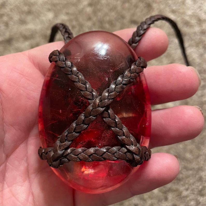 May include: A red, oval-shaped pendant with a brown leather cord wrapped around it. The pendant is translucent and has a slightly cloudy appearance.