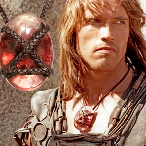 May include: A close-up of a red gemstone pendant with a leather cord. The gemstone is oval-shaped and has a dark red color with a slight shimmer. The leather cord is braided and wraps around the gemstone.